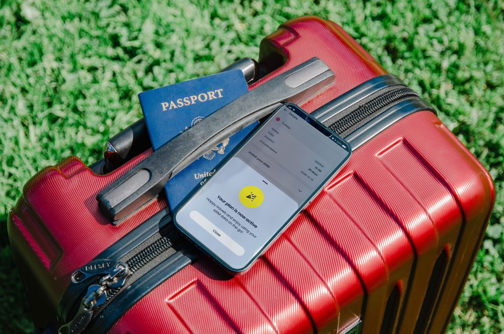 Smartphone displaying “Your plan is now active” screen rests on a red suitcase beside a U.S. passport, representing easy travel with Saily eSIM.
